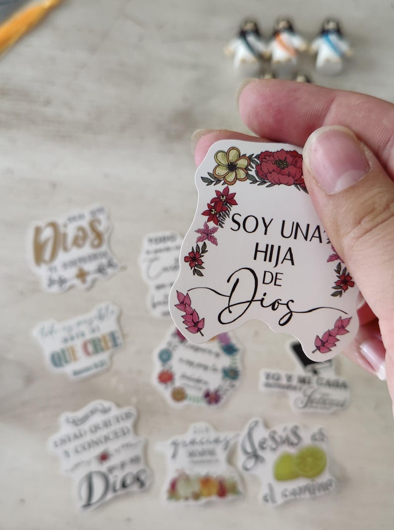May include: A collection of stickers with floral designs and religious phrases in Spanish. One sticker reads "SOY UNA HIJA DE DIOS" with a floral border. Other stickers feature phrases like "Dios" and "QUE CREE".