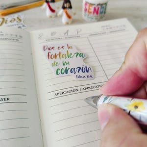 May include: A notebook with lined pages, a pen, and a sticker with the text "Dios es la fortaleza de mi coraz&oacute;n" in colorful lettering. Two small figurines and a candle with the word "SEND" are also visible.