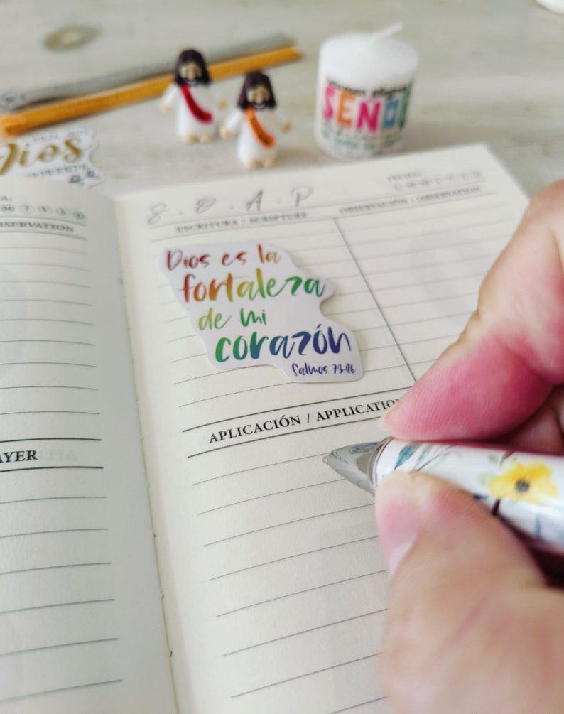 May include: A notebook with lined pages, a pen, and a sticker with the text "Dios es la fortaleza de mi coraz&oacute;n" in colorful lettering. Two small figurines and a candle with the word "SEND" are also visible.