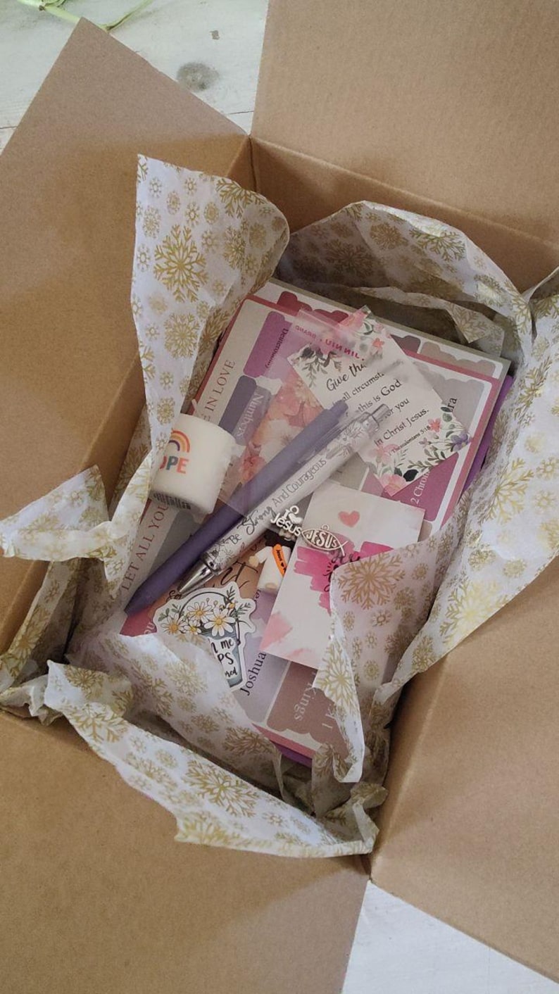 May include: A cardboard box filled with stationery items, including pens, stickers, and cards. The box contains a pen with the text "Be Strong And Courageous" and a sticker with the word "HOPE". The box is lined with tissue paper.