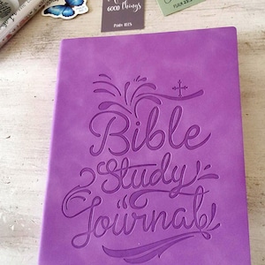 May include: A purple Bible Study Journal with embossed lettering. The journal has a ribbon bookmark. Also pictured are decorative bookmarks with floral designs and religious quotes, and small figurines.