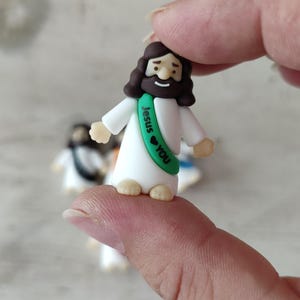 May include: A small figurine of Jesus wearing a white robe and a green sash with the words "Jesus ❤ You." The figurine has brown hair and a beard. Other similar figurines are slightly out of focus in the background.