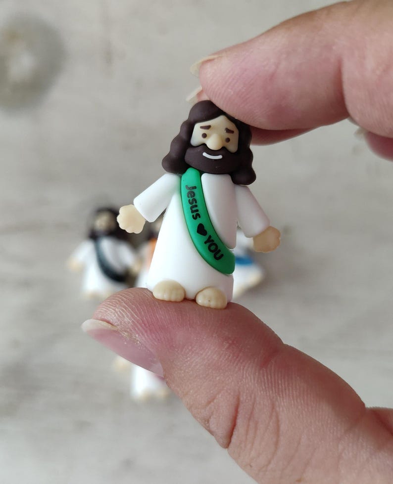 May include: A small figurine of Jesus wearing a white robe and a green sash with the words "Jesus ❤ You." The figurine has brown hair and a beard. Other similar figurines are slightly out of focus in the background.