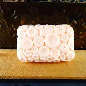 May include: A pink soap bar shaped like a button with a button design. The soap is made to look like a collection of buttons.