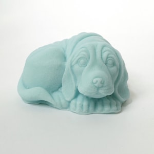 May include: A light blue ceramic sculpture of a dog lying down. The dog has a detailed face with brown eyes and a black nose.
