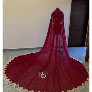 Può includere: Un lungo velo trasparente rosso scuro con piccole decorazioni bianche sparse e un ampio bordo smerlato ricamato in oro. Il tessuto cade a terra in uno strascico circolare, con le lettere AS ricamate in bianco vicino al bordo inferiore.