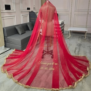 Pode incluir: Um véu de noiva vermelho vibrante com acabamento dourado e lantejoulas espalhadas é drapeado sobre um manequim. O véu apresenta o texto bordado "Forever you & me 25.02.2026". O véu é exibido em uma sala bem iluminada com um sofá cinza e móveis brancos.