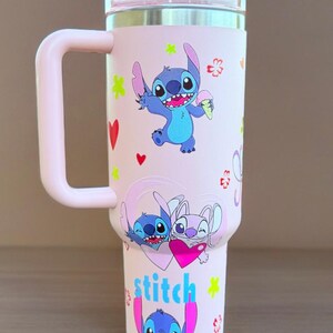 May include: A pink insulated travel mug decorated with Stitch from Lilo & Stitch. The mug has a pink handle and lid, with various images of Stitch and floral designs. The word "stitch" is written in blue and pink.