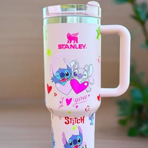May include: A pink Stanley travel mug with a cartoon image of Stitch and another character holding a heart. The mug has a handle and a lid. The text "I love you" and "Stitch" are also on the mug.