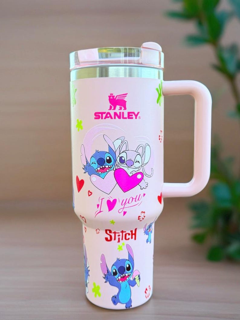 May include: A pink Stanley travel mug with a cartoon image of Stitch and another character holding a heart. The mug has a handle and a lid. The text "I love you" and "Stitch" are also on the mug.