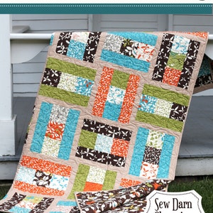 May include: A colorful quilt with a patchwork design featuring brown, green, orange, blue, and white fabrics. The quilt is folded and lying on a grassy surface. The text "Sew Darn Charming by Janel Mettsker" is visible on the quilt.