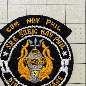 May include: A circular embroidered patch with a black background and gold text reading COM NAV PHIL, S.R.F. SUBIC BAY PHIL, and DIVING SALVAGE. The center features a silver diving helmet, a trident, and two gold dolphins with a yellow 30 sticker.