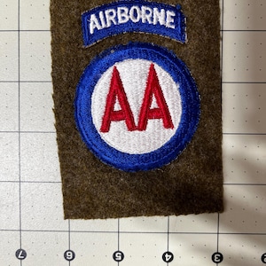May include: A rectangular olive drab felt patch featuring an embroidered blue and white 'AIRBORNE' tab above a circular patch with a white center, red 'AA' lettering, and a thick blue border, placed on a grid mat measuring 5 inches wide.