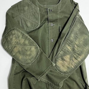 May include: An olive green shooting jacket featuring a round neckline, a front button closure, and reinforced padded patches on the shoulders and elbows. The fabric shows signs of wear with faded, lighter-colored areas on the elbow pads.