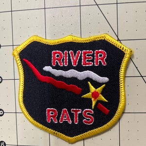 May include: A shield-shaped embroidered patch with a black background and a yellow border. The text RIVER RATS is stitched in red with white outlines. It features a wavy white line, a red wavy line, and a yellow star with a red center and handle.