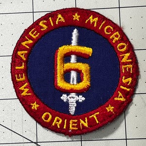 May include: A circular embroidered patch featuring a dark blue center with a silver sword and a large yellow number 6. The red border contains the yellow embroidered text MELANESIA, MICRONESIA, and ORIENT, separated by small yellow stars.