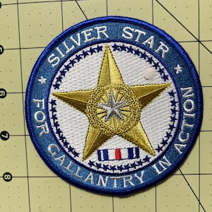 May include: A circular embroidered patch featuring a large gold star with a smaller silver star at its center, set against a white background. The blue border contains the text SILVER STAR FOR GALLANTRY IN ACTION and a small ribbon graphic.