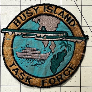May include: A circular embroidered patch featuring a light blue background with a tan border. The text BUSY ISLAND TASK FORCE is stitched in black. The design includes a gray ship, a green palm tree, a landmass, and a dark green airplane.