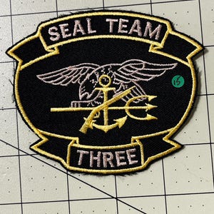 May include: A black embroidered patch featuring the text SEAL TEAM at the top and THREE at the bottom in light-colored thread. The center displays a gold anchor and trident emblem overlaid with a light-colored eagle graphic on a black background.