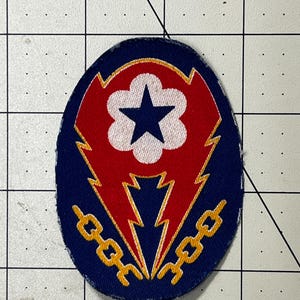 May include: An oval-shaped embroidered patch featuring a dark blue background with a central red lightning bolt shape. Inside the red shape is a white flower-like emblem containing a dark blue star. Two yellow chain links flank the bottom.