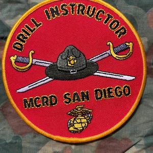 May include: A circular embroidered patch with a red background and a yellow border. It features the text DRILL INSTRUCTOR and MCRD SAN DIEGO in black. The center displays a black campaign hat over two crossed swords and a gold eagle, globe, and anchor.