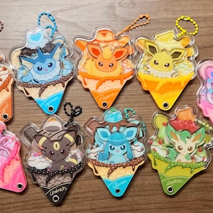 Puede incluir: Una colección de diez colgantes acrílicos transparentes con varios personajes de Pokémon diseñados como conos de helado. Cada colgante tiene un personaje diferente, un esquema de color y una pequeña cadena. Dispuestos sobre una superficie de madera.