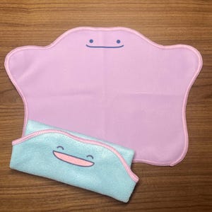 Könnte beinhalten: Ein hellviolettes, amorphes Mikrofasertuch mit einer einfachen dunkelblauen Linie als Mund und zwei Punkten als Augen. Ein kleineres, gefaltetes hellblaues Tuch mit rosa Rand und einer lächelnden Gesichtsgrafik befindet sich im Vordergrund.