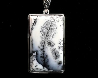 May include: A rectangular pendant with a silver frame and a white stone featuring black dendritic inclusions, suspended from a silver chain. The stone's pattern resembles a tree or landscape. The pendant is displayed against a black background.