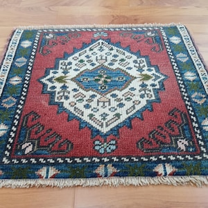 May include: A small, square rug featuring a central geometric pattern in white, blue, and green on a red field. It has a blue border with repeating motifs and a fringed edge. The rug is displayed on a wooden floor.