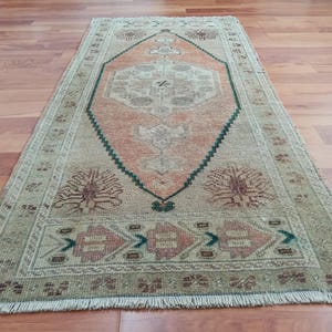 May include: Rectangular rug with a central diamond-shaped design in peach and beige, framed by a dark green border. The rug features intricate floral and geometric patterns in shades of brown, green, and peach. The rug is placed on a wooden floor.