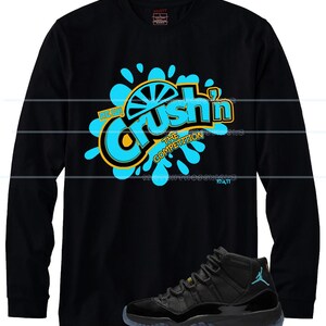 Puede incluir: Camiseta negra de manga larga con la frase "Crush'n The Competition" en azul y amarillo. Un par de zapatillas deportivas negras y azules se encuentran en la parte inferior de la imagen.