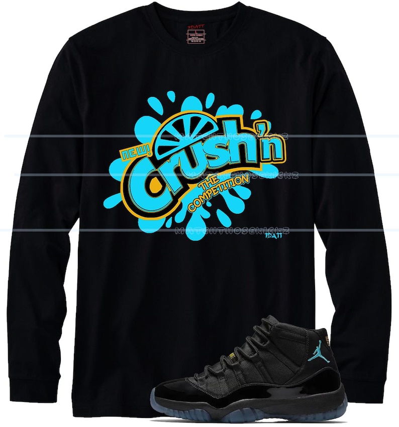 Puede incluir: Camiseta negra de manga larga con la frase "Crush'n The Competition" en azul y amarillo. Un par de zapatillas deportivas negras y azules se encuentran en la parte inferior de la imagen.