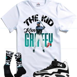 May include: A white short-sleeve t-shirt featuring a graphic of Ken Griffey Jr. batting with the text THE KID and KEN GRIFFEY JR. in black and teal, a pair of black and white patterned crew socks, and a black and white athletic sneaker with the number 24.