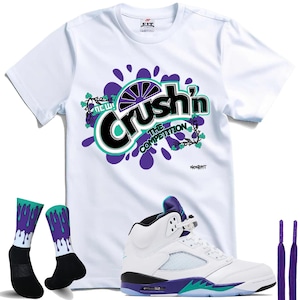 May include: White t-shirt with "Crush'n The Competition" graphic, matching socks, white and purple sneakers, and purple laces. The shirt features a colorful design with purple and teal accents. The sneakers have a white upper with purple and teal details.