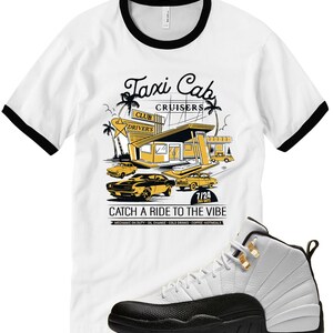 May include: White t-shirt with black trim featuring a yellow and black graphic of a retro taxi cab service. The graphic includes the text "Taxi Cab Cruisers" and "Catch a Ride to the Vibe". A white and black sneaker is in the foreground.