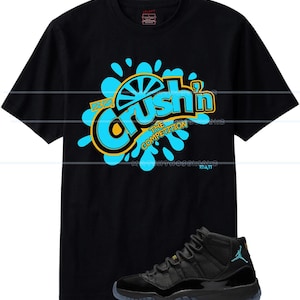 Puede incluir: Camiseta negra con un gr&aacute;fico que dice "Crush'n The Competition" en azul y amarillo. Un par de zapatillas negras y azules est&aacute;n en primer plano. Las zapatillas tienen un logotipo Jumpman azul.