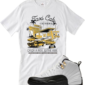May include: White t-shirt with a graphic of a retro taxi cab scene, including cars and a building with the text "Taxi Cab Cruisers." Paired with white and black sneakers with gold accents. The shirt also has the text "Catch a Ride to the Vibe."