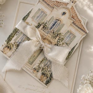Op de afbeelding: Een uitnodigingskaart met een aquarel illustratie van een huis, vastgebonden met een crèmekleurig lint. De kaart luidt: "U bent uitgenodigd voor een speciale viering."