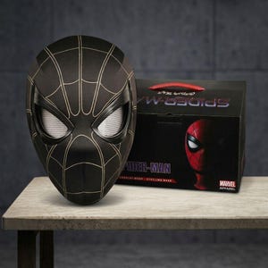May include: A black Spider-Man mask with gold web detailing and white mesh eye lenses, accompanied by a black product box featuring the text Spider-Man, Marvel Apparel, and a graphic of a red Spider-Man mask on the side of the packaging.