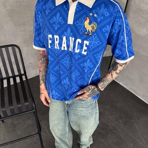 Peut inclure: Un polo bleu &agrave; manches courtes avec un col blanc, un motif r&eacute;p&eacute;t&eacute; de fleurs de lys et le mot FRANCE imprim&eacute; en blanc. Un embl&egrave;me de coq dor&eacute; avec deux petites &eacute;toiles dor&eacute;es est plac&eacute; au-dessus du texte. Le polo pr&eacute;sente des d&eacute;tails passepoil&eacute;s blancs.