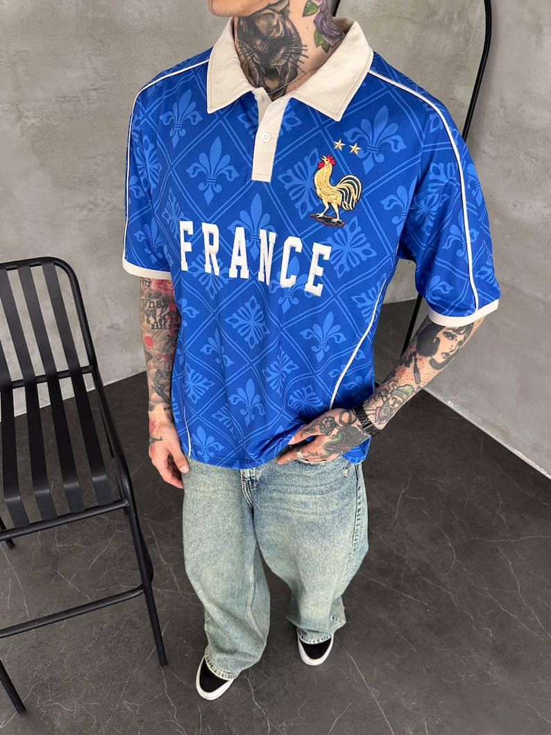 Peut inclure: Un polo bleu &agrave; manches courtes avec un col blanc, un motif r&eacute;p&eacute;t&eacute; de fleurs de lys et le mot FRANCE imprim&eacute; en blanc. Un embl&egrave;me de coq dor&eacute; avec deux petites &eacute;toiles dor&eacute;es est plac&eacute; au-dessus du texte. Le polo pr&eacute;sente des d&eacute;tails passepoil&eacute;s blancs.