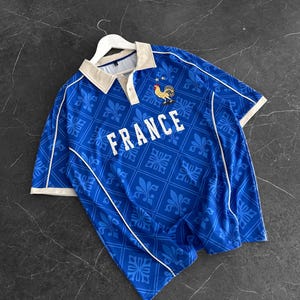 Peut inclure: Un polo bleu &agrave; manches courtes avec un motif r&eacute;p&eacute;t&eacute; de fleurs de lys, un col beige et un passepoil blanc. La poitrine affiche le mot FRANCE en lettres capitales blanches au-dessus d'un embl&egrave;me de coq dor&eacute; et bleu avec deux petites &eacute;toiles.