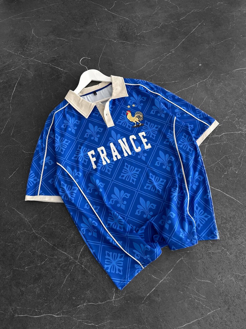 Peut inclure: Un polo bleu &agrave; manches courtes avec un motif r&eacute;p&eacute;t&eacute; de fleurs de lys, un col beige et un passepoil blanc. La poitrine affiche le mot FRANCE en lettres capitales blanches au-dessus d'un embl&egrave;me de coq dor&eacute; et bleu avec deux petites &eacute;toiles.