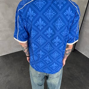Peut inclure: Un polo bleu &agrave; manches courtes orn&eacute; d'un motif r&eacute;p&eacute;titif en losanges avec des symboles de fleur de lys. Le v&ecirc;tement pr&eacute;sente un col blanc et des bordures blanches aux manches, ainsi que des d&eacute;tails de passepoil blanc sur les &eacute;paules.