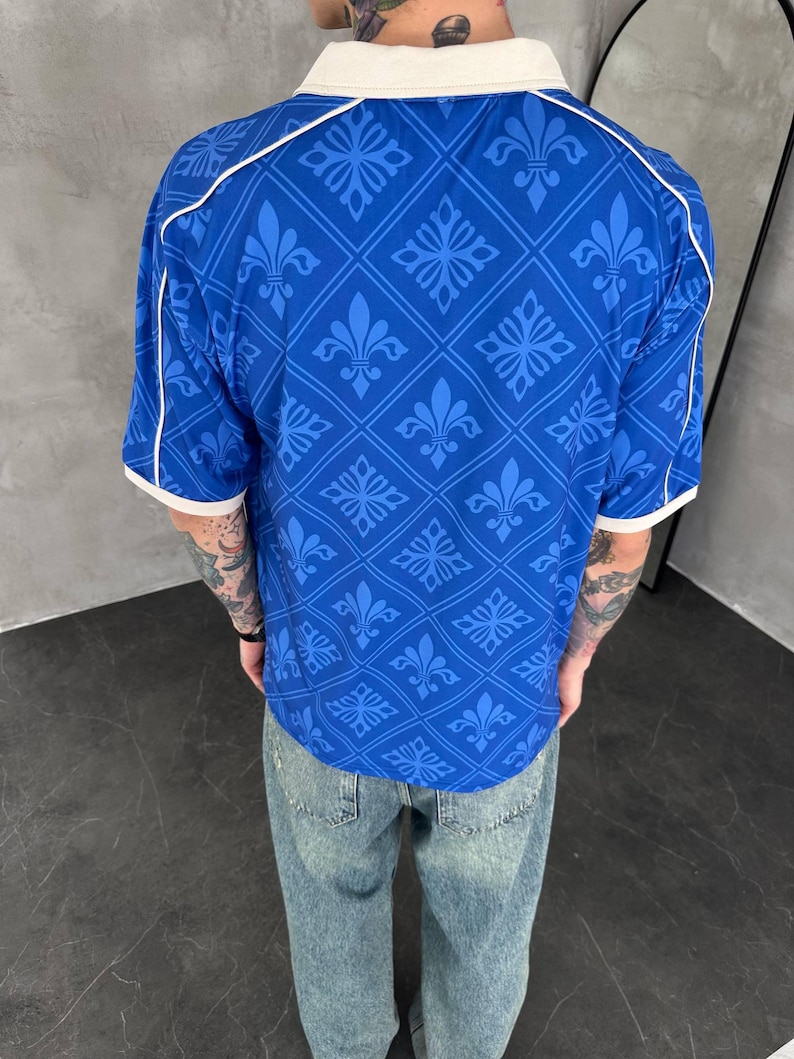Peut inclure: Un polo bleu &agrave; manches courtes orn&eacute; d'un motif r&eacute;p&eacute;titif en losanges avec des symboles de fleur de lys. Le v&ecirc;tement pr&eacute;sente un col blanc et des bordures blanches aux manches, ainsi que des d&eacute;tails de passepoil blanc sur les &eacute;paules.