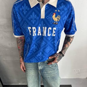 Peut inclure: Un polo bleu avec un col blanc et des bordures blanches sur les manches. Le v&ecirc;tement pr&eacute;sente un motif r&eacute;p&eacute;titif de fleurs de lys, le mot FRANCE en lettres blanches et un embl&egrave;me de coq dor&eacute; avec deux petites &eacute;toiles dor&eacute;es sur la poitrine.