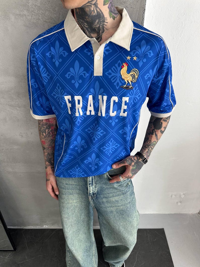 Peut inclure: Un polo bleu avec un col blanc et des bordures blanches sur les manches. Le v&ecirc;tement pr&eacute;sente un motif r&eacute;p&eacute;titif de fleurs de lys, le mot FRANCE en lettres blanches et un embl&egrave;me de coq dor&eacute; avec deux petites &eacute;toiles dor&eacute;es sur la poitrine.
