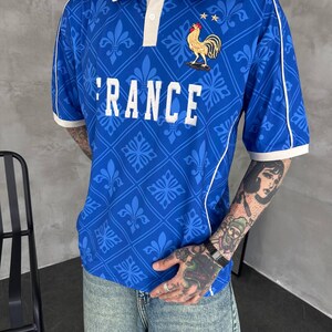Peut inclure: Un polo bleu avec un col blanc, des passepoils blancs et un motif r&eacute;p&eacute;titif de fleurs de lys. Le v&ecirc;tement pr&eacute;sente le texte RANCE en lettres blanches et un embl&egrave;me de coq dor&eacute; brod&eacute; surmont&eacute; de deux petites &eacute;toiles sur la poitrine.