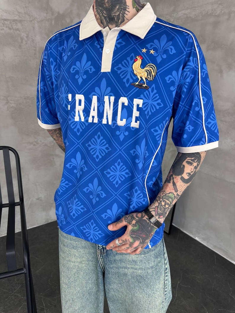 Peut inclure: Un polo bleu avec un col blanc, des passepoils blancs et un motif r&eacute;p&eacute;titif de fleurs de lys. Le v&ecirc;tement pr&eacute;sente le texte RANCE en lettres blanches et un embl&egrave;me de coq dor&eacute; brod&eacute; surmont&eacute; de deux petites &eacute;toiles sur la poitrine.
