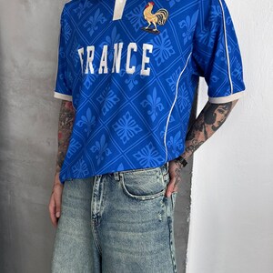 Peut inclure: Un polo bleu &agrave; manches courtes avec un col blanc, des passepoils blancs et un motif r&eacute;p&eacute;t&eacute; de fleurs de lys. Le mot FRANCE est imprim&eacute; en blanc sur la poitrine, accompagn&eacute; d'un embl&egrave;me de coq dor&eacute; et de deux petites &eacute;toiles dor&eacute;es au-dessus.
